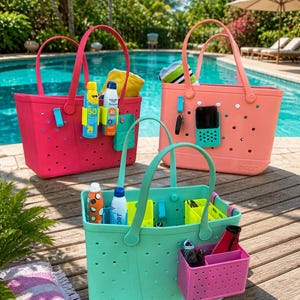 May include: Three colorful tote bags in pink, coral, and mint green sit on a wooden deck near a pool. Each bag has a matching handle and a variety of accessories, including sunscreen, keys, and a phone. The bags are designed for beach or pool use.