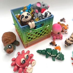 May include: A rainbow-colored plastic crate filled with small 3D-printed animal figures including a sloth, a mouse, and a raccoon wearing a Santa hat. Surrounding the crate are various articulated figures: a brown turtle, a red crab, and a green lizard.