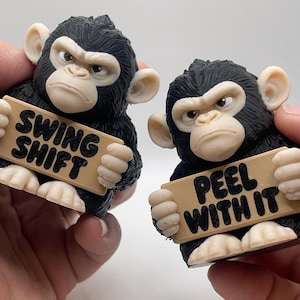 May include: Two small black and beige chimpanzee figurines with textured fur. One holds a sign reading SWING SHIFT, and the other holds a sign reading PEEL WITH IT. Both figures have light-colored faces, ears, and hands with a serious expression.