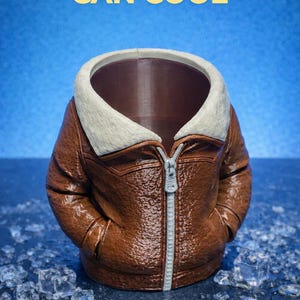 May include: A brown can cooler shaped like an aviator bomber jacket with a white faux fur collar and a silver zipper. The text "MAKE YOUR CAN COOL" is at the top, and "Aviator Bomber Jacket Can Cooler" is at the bottom.