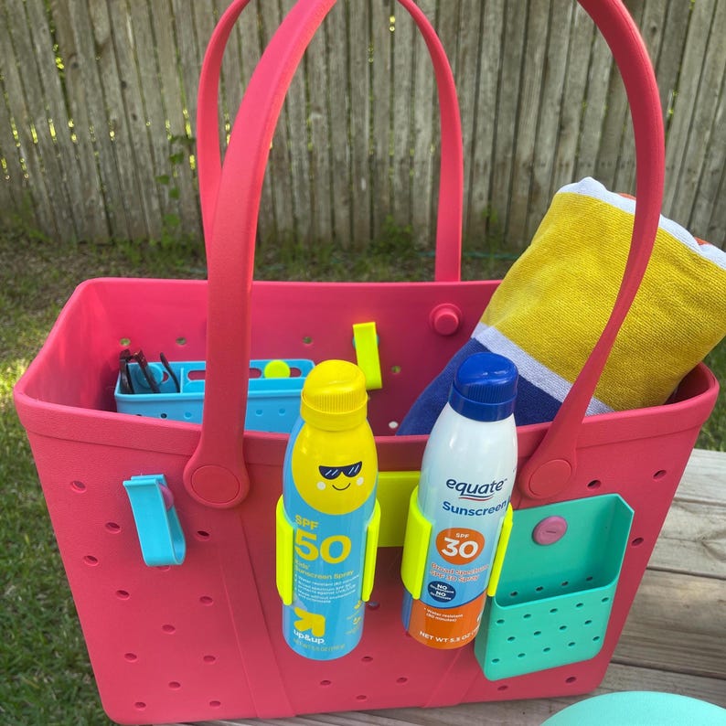 May include: A bright pink tote bag with matching handles, holding sunscreen, a beach towel, and small blue and green organizers. The bag has a perforated design and is ready for a day at the beach.