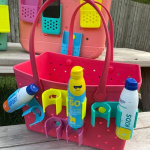 May include: A pink tote bag with a matching handle, featuring various colored accessories. The bag holds sunscreen bottles and has attached holders for phones and other items. The bag is designed for beach or outdoor use.