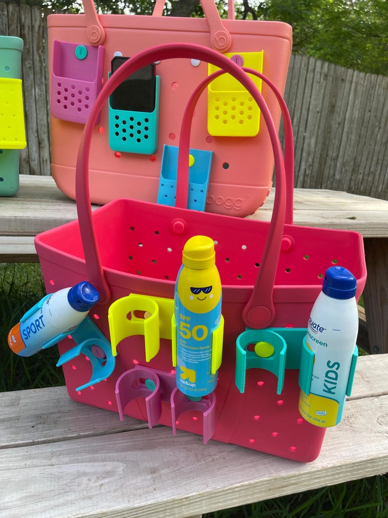 May include: A pink tote bag with a matching handle, featuring various colored accessories. The bag holds sunscreen bottles and has attached holders for phones and other items. The bag is designed for beach or outdoor use.
