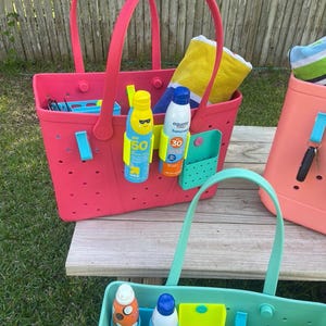 May include: A collection of colorful tote bags, perfect for the beach or pool. The bags are pink, coral, and teal, with matching handles. Each bag contains sunscreen, towels, and other beach essentials. The bags have multiple pockets and are made of a durable material.