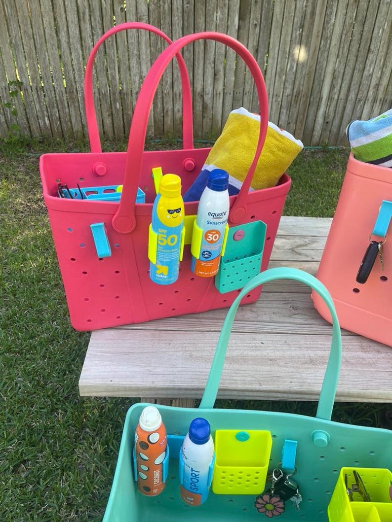 May include: A collection of colorful tote bags, perfect for the beach or pool. The bags are pink, coral, and teal, with matching handles. Each bag contains sunscreen, towels, and other beach essentials. The bags have multiple pockets and are made of a durable material.