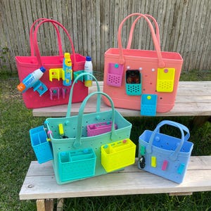 May include: A collection of colorful tote bags in pink, coral, mint green, and blue, each with matching accessories. The bags have handles and are designed with multiple compartments and attachments for organizing items. Some bags have accessories like sunscreen and water bottles.