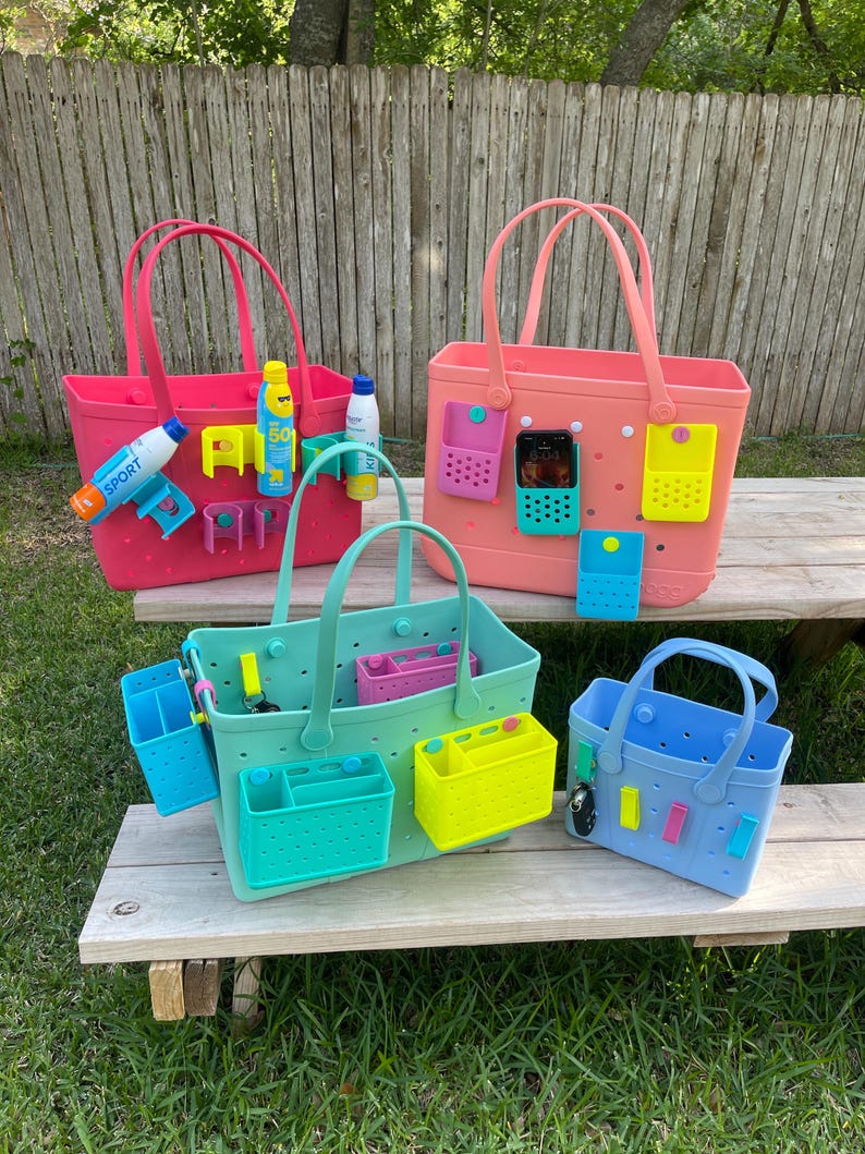 May include: A collection of colorful tote bags in pink, coral, mint green, and blue, each with matching accessories. The bags have handles and are designed with multiple compartments and attachments for organizing items. Some bags have accessories like sunscreen and water bottles.