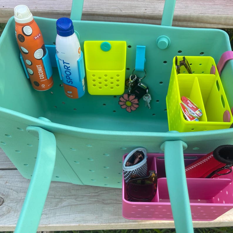 May include: A teal tote bag with various accessories. Inside are two bottles, one orange with "Coconut" text and one white with "Sport" text. There are also yellow and pink organizers, keys, sunglasses, and a red item.