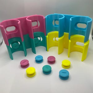 May include: A set of colorful plastic toy building blocks in pink, teal, yellow, and blue. The blocks have a curved design with a handle-like opening at the top. Several small, round, two-toned pieces in matching colors are also included.