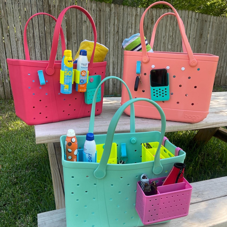 May include: Three colorful tote bags in pink, coral, and teal, each with matching handles. The bags are filled with beach essentials like sunscreen, towels, and accessories. The bags have a perforated design and are sitting on a wooden picnic table.