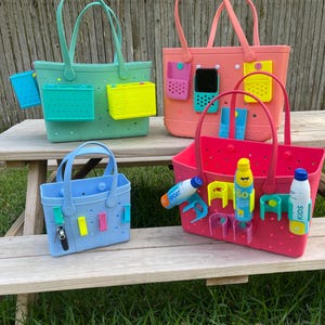 May include: Four brightly coloured tote bags with handles and various attached accessories. The bags are in shades of blue, green, pink, and red. Each bag has a different set of accessories, including small containers and phone holders. The bags are on a wooden table.