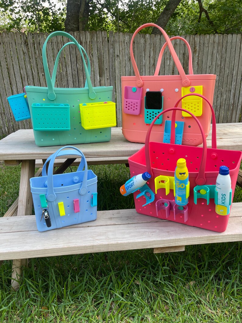 May include: Four brightly coloured tote bags with handles and various attached accessories. The bags are in shades of blue, green, pink, and red. Each bag has a different set of accessories, including small containers and phone holders. The bags are on a wooden table.