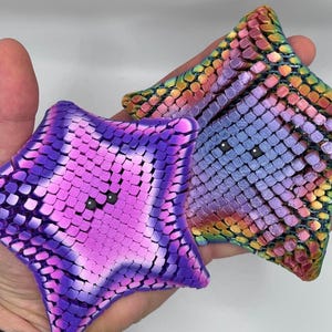 May include: Two star-shaped objects with a textured, mosaic-like surface. One star features a gradient of pink and purple tones, while the other displays a multi-colored iridescent pattern of blue, purple, yellow, and orange, both with small eyes.