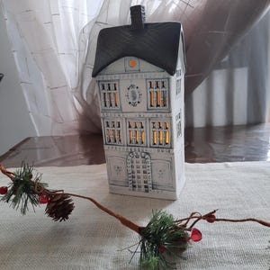Könnte beinhalten: Ein weißes Keramik-Haus-Teelichthalter mit schwarzem Dach und Schornstein. Die Fenster sind mit einem warmen Schein beleuchtet. Ein dekorativer Zweig mit Tannennadeln, roten Beeren und einem Tannenzapfen befindet sich im Vordergrund.