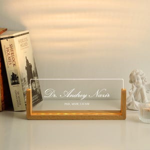 May include: A lighted desk nameplate with the text "Dr. Andrey Nazir, PhD, MSW, LICSW" in elegant script. The nameplate is made of clear acrylic and sits in a wooden base. Books and a small angel statue are in the background.