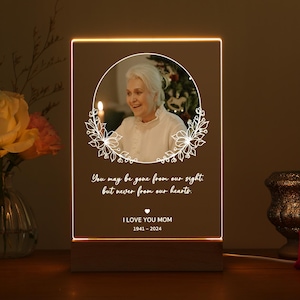 Puede incluir: Placa conmemorativa iluminada con la foto de una mujer dentro de una corona floral. El texto dice "You may be gone from our sight but never from our hearts. I LOVE YOU MOM 1941-2024". La placa está sobre una base de madera.