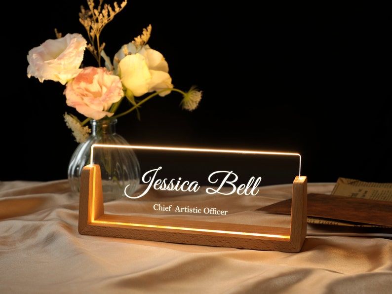 May include: A wooden desk sign with a clear acrylic insert. The sign is illuminated with warm white LED lights. The text on the sign reads "Jessica Bell, Chief Artistic Officer".