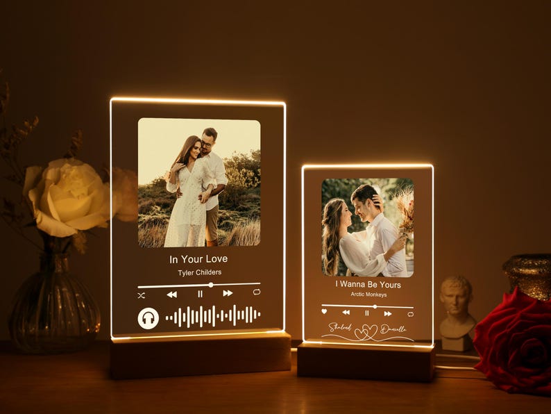 Puede incluir: Dos marcos de fotos iluminados con fotos de parejas y t&iacute;tulos de canciones. El marco superior presenta una pareja en un campo con el t&iacute;tulo de la canci&oacute;n "In Your Love" de Tyler Childers. El marco inferior presenta una pareja abraz&aacute;ndose con el t&iacute;tulo de la canci&oacute;n "I Wanna Be Yours" de Arctic Monkeys.
