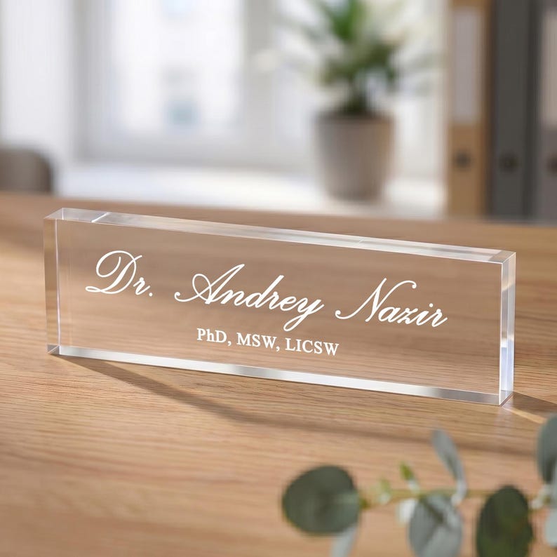 May include: A clear acrylic desk nameplate with the name "Dr. Andrey Nazir" in elegant white script. The nameplate also includes the credentials "PhD, MSW, LICSW" in a smaller font. The nameplate is rectangular and sits on a wooden surface.