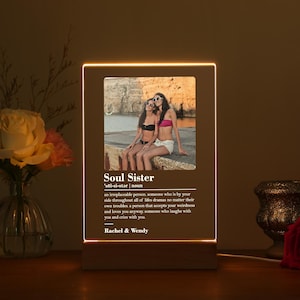 Könnte beinhalten: Ein beleuchteter Fotorahmen mit einem Bild von zwei Frauen in Badeanzügen am Strand. Der Rahmen hat einen weißen Hintergrund mit dem Text "Soul Sister" und einer Definition des Wortes. Die Namen "Rachel & Wendy" sind unten auf dem Rahmen gedruckt.