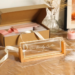 May include: A personalized desk nameplate with the text "Dr. Emily Carter" in elegant script, illuminated by LED lights. The nameplate is set in a wooden base and comes with a gift box and ribbon.