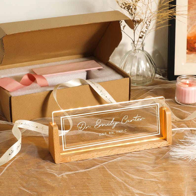 May include: A personalized desk nameplate with the text "Dr. Emily Carter" in elegant script, illuminated by LED lights. The nameplate is set in a wooden base and comes with a gift box and ribbon.
