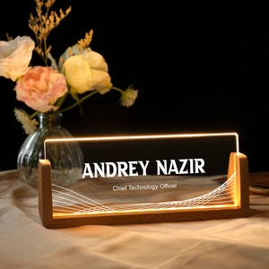 May include: A wooden desk nameplate with a clear acrylic insert. The name "ANDREY NAZIR" is printed in white on the acrylic insert, along with the title "Chief Technology Officer". The nameplate is illuminated from behind with warm white LED lights.