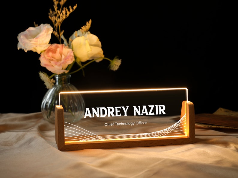 May include: A wooden desk nameplate with a clear acrylic insert. The name "ANDREY NAZIR" is printed in white on the acrylic insert, along with the title "Chief Technology Officer". The nameplate is illuminated from behind with warm white LED lights.