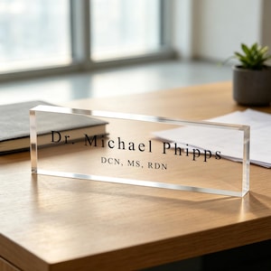 May include: A clear acrylic desk nameplate with the name "Dr. Michael Phipps" and credentials printed in black. The nameplate is rectangular and sits on a wooden desk, with a notebook and plant in the background.