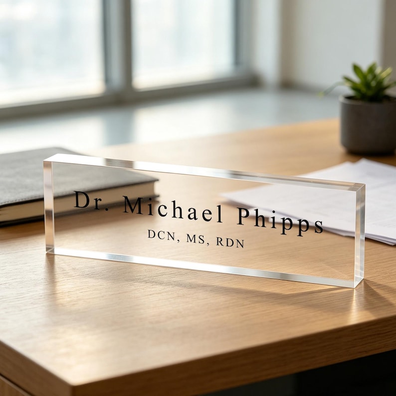 May include: A clear acrylic desk nameplate with the name "Dr. Michael Phipps" and credentials printed in black. The nameplate is rectangular and sits on a wooden desk, with a notebook and plant in the background.