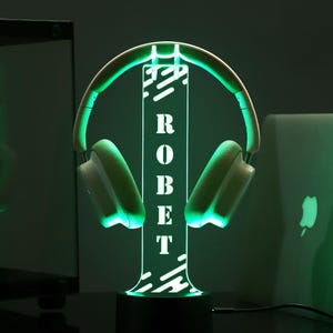 Puede incluir: Un soporte de auriculares con iluminaci&oacute;n verde con el nombre "ROBERT" en vertical. El soporte sostiene unos auriculares blancos y verdes. La base es negra y el fondo es oscuro.