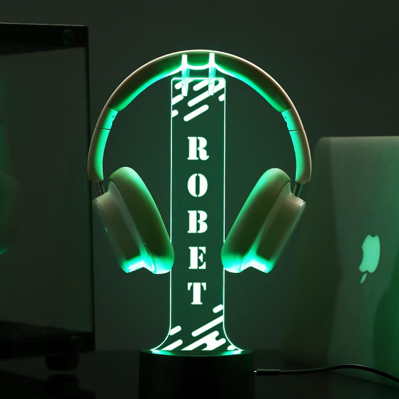 Puede incluir: Un soporte de auriculares con iluminaci&oacute;n verde con el nombre "ROBERT" en vertical. El soporte sostiene unos auriculares blancos y verdes. La base es negra y el fondo es oscuro.