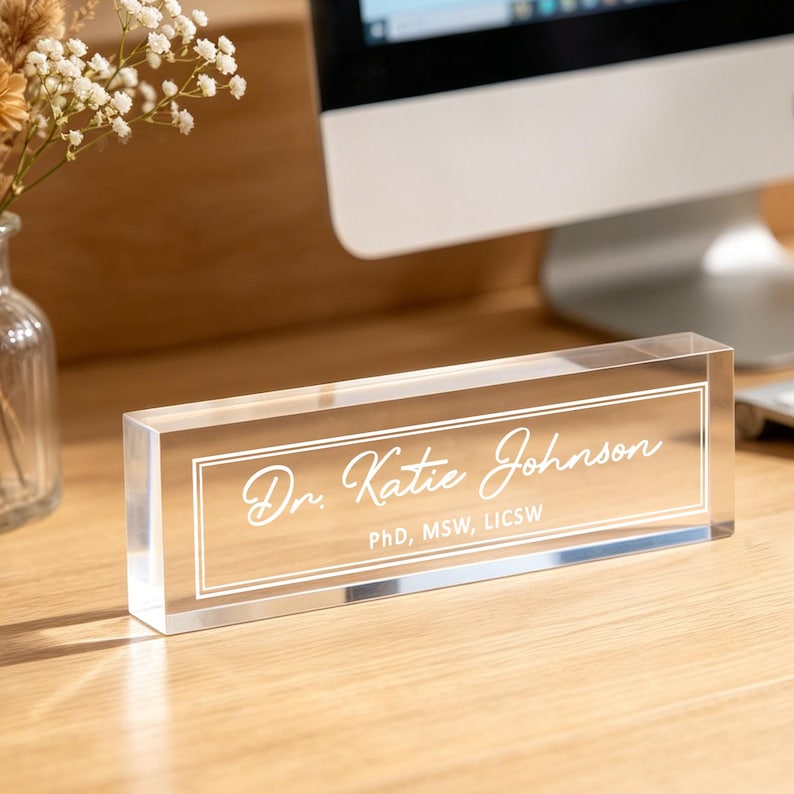 May include: A clear acrylic desk nameplate with the name "Dr. Katie Johnson" in elegant white script. The nameplate also includes the credentials "PhD, MSW, LICSW" in a smaller font, all within a white border.