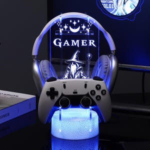 May include: A gaming headset stand with a controller base and a glowing acrylic panel. The panel features the word "GAMER" and an illustration of a wizard. The stand is illuminated with blue light.