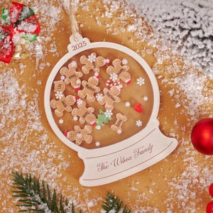 Könnte beinhalten: Ein weißer, schneekugel-förmiger Weihnachtsschmuck mit Lebkuchenmännchen im Inneren. Der Schmuck trägt das Jahr 2025 und den Familiennamen "The Wilson Family". Die Lebkuchenmännchen tragen Namen.