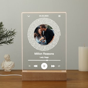 May include: A white LED light with a wooden base displays a song title, artist, and date. The song is "Million Reasons" by Lady Gaga. A photo of a couple is displayed in a circle in the center of the light.