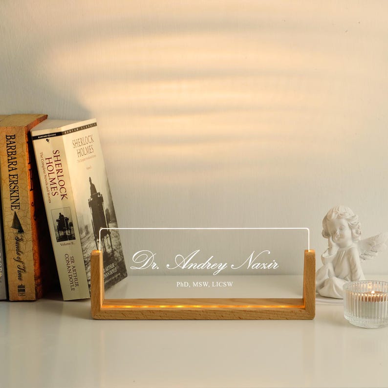 May include: A wooden-based illuminated nameplate with the text "Dr. Andrey Nazir, PhD, MSW, LICSW" in a stylish script. The nameplate is made of clear acrylic. Books and a small angel statue are in the background.