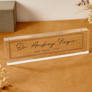 May include: A clear acrylic desk nameplate with the name "Dr. Andrey Nazin" in elegant script. The nameplate also includes the credentials "PhD, MSW, LICSW" in a smaller font. The background is a light brown color.