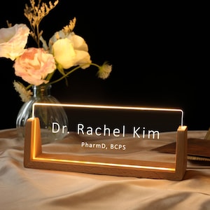 May include: A wooden desk nameplate with the name "Dr. Rachel Kim" and credentials "PharmD, BCPS" illuminated by a warm light. The nameplate is set on a wooden base. A vase of flowers is in the background.