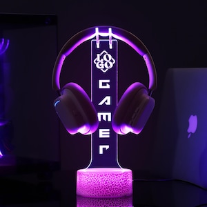 May include: A purple LED headphone stand with the word "GAMER" vertically displayed. The stand holds a pair of white and purple headphones. The base of the stand has a textured pattern, and the entire setup glows with purple light.