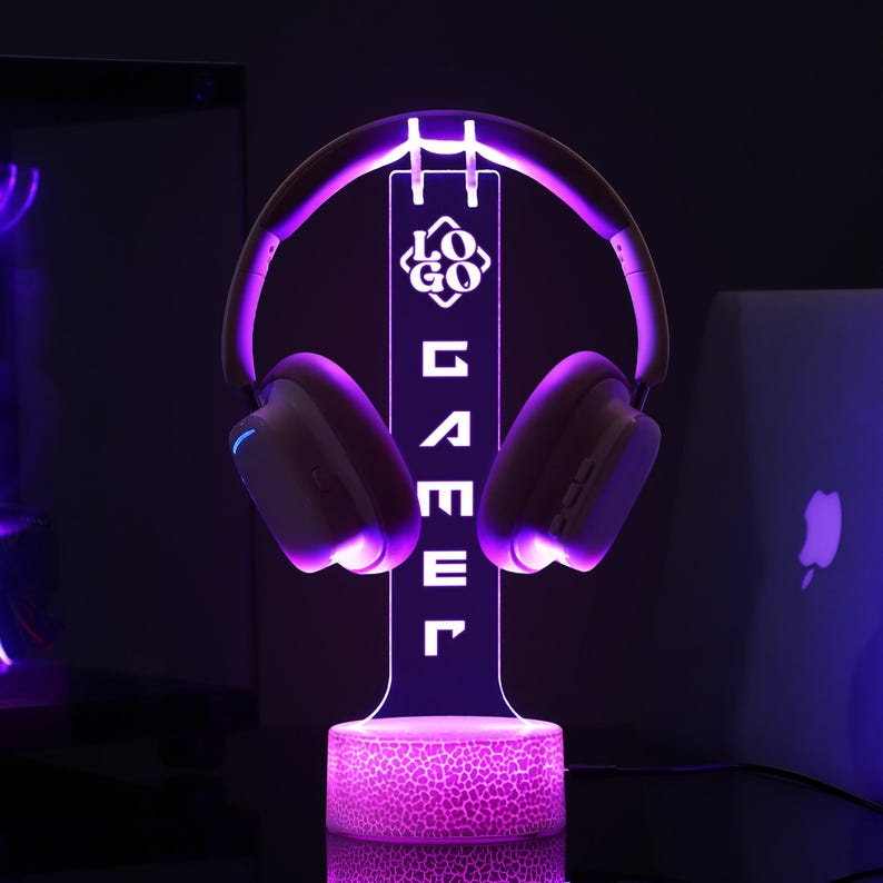 Puede incluir: Un soporte para auriculares LED p&uacute;rpura con la palabra "GAMER" mostrada verticalmente. El soporte sostiene un par de auriculares blancos y p&uacute;rpuras. La base del soporte tiene un patr&oacute;n texturizado y brilla en p&uacute;rpura. Un logotipo se muestra encima de la palabra "GAMER".