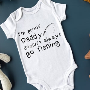 Puede incluir: Un body blanco para bebé con texto negro que dice "Soy la prueba de que papá no siempre va a pescar" y un gráfico de caña de pescar negra.