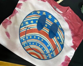 Puede incluir: Una camiseta blanca con un patrón de tie-dye rojo, blanco y azul. La camiseta presenta un gran gráfico de cara sonriente con un diseño de bandera estadounidense.