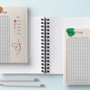 以下が含まれることがあります： グリッドページのある2冊のリング綴じジャーナル。1冊のジャーナルの表紙には「Rate My Day」と書かれており、評価用のカラーコードキーが含まれています。もう1冊のジャーナルの表紙には「Health Log」と書かれており、こちらもカラーコードキーが付いています。2本の鉛筆が前景にあります。