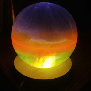 May include: A glowing, spherical lamp with a gradient of colors. The top is a deep purple, transitioning to orange, yellow, and green at the base. It sits on a round, wooden base, creating a warm, ambient light.