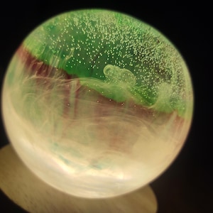 May include: A spherical glass paperweight with an internal design. The top half is green with tiny white bubbles, transitioning to a maroon band, and then a swirling white and clear base. The paperweight is illuminated from below, creating a glowing effect.