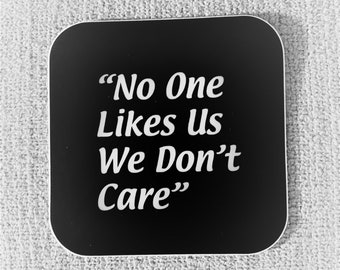 May include: Black square coaster with white text that reads "No One Likes Us We Don't Care"