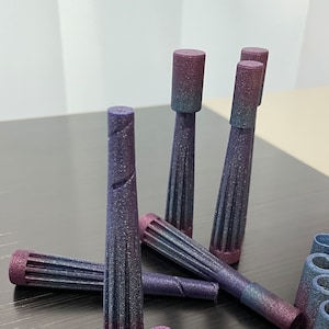 Puede incluir: Un grupo de objetos cilíndricos de color púrpura y azul iridiscente, con textura. Algunos de los objetos están de pie, mientras que otros están tumbados sobre una superficie oscura.