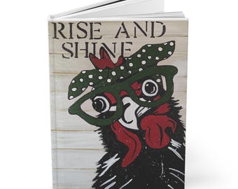 May include: A white journal with a black and green illustration of a rooster wearing glasses and a polka dot bandana. The text "Rise and Shine" is printed on the cover.