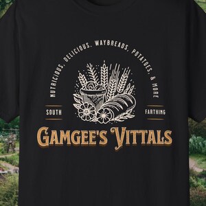 Op de afbeelding: Een zwart t-shirt met korte mouwen en een witte en gouden grafische print. Het ontwerp bevat illustraties van tarwe, een mand en brood, met de tekst "NUTRITIOUS, DELICIOUS, WAYBREADS, POTATOES, & MORE" en "SOUTH FARTHING GAMGEE'S VITTALS".